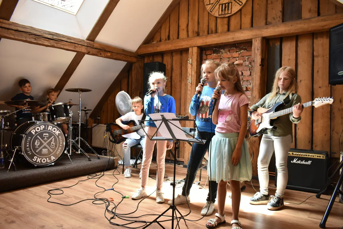 Bērnu grupa mēģina koka bēniņu studijā: trīs dziedātājas, ģitāra, basģitāra un bungas
